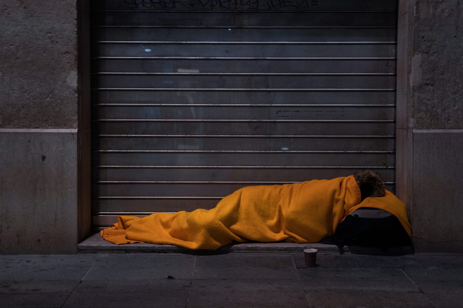 a-homeless-person-sleeps-on-the-street-under-a-blanket-in-front-of-a-storefront